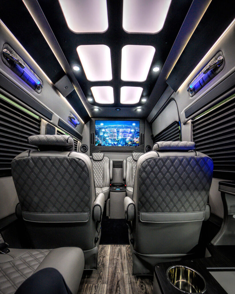Interior photo of Mercedes-Benz Sprinter Van luxury limo conversion, facing toward the front of the vehicle and showing one of the HDTVs, as well as the leather captains chairs